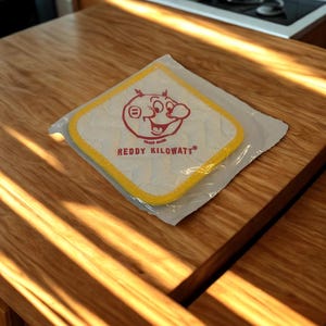 Op de afbeelding: Een vierkante witte doorgestikte pannenlap met een gele stoffen rand, verpakt in doorzichtig plastic. Het midden toont een rode afbeelding van het personage Reddy Kilowatt met de tekst REDDY KILOWATT in het rood onder de illustratie.