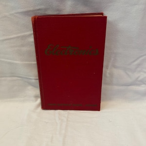 May include: A red book titled "Electronics" in green script. The book has a hard cover and appears to be vintage. The title is centered on the front cover. The book is on a white surface.