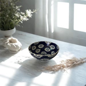 Puede incluir: Cuenco de cerámica decorativo con fondo azul oscuro y estampado floral blanco. El cuenco tiene un borde dorado y está sobre una mesa blanca. La imagen también incluye un jarrón con flores blancas y un elemento decorativo.