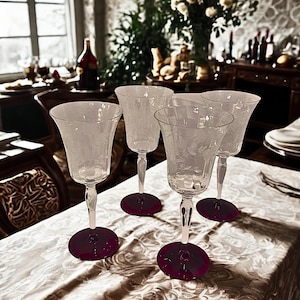 May include: Four clear wine glasses with etched floral patterns, each with a stem and a deep purple base, are arranged on a white tablecloth with a floral design. The glasses are in focus, suggesting a formal dining setting.