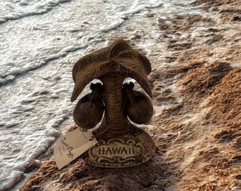 Könnte beinhalten: Braune, dekorative Palmenfigur mit Kokosnüssen, auf einem Sandstrand mit Meereswellen im Hintergrund. Auf der Basis der Figur steht "HAWAII". Ein Etikett trägt die Aufschrift "Treasure Craft".