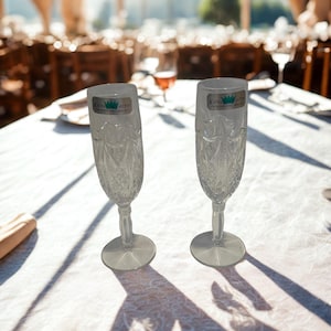 May include: Two clear crystal champagne flutes with intricate cut glass patterns. Each glass has a round base and a long stem. The flutes are on a white tablecloth, with the text "KATHA HUTTE" visible on the glasses.