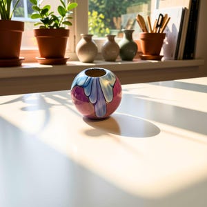 May include: A spherical ceramic vase with a glossy finish, featuring a deep magenta base color and a decorative light blue drip pattern cascading from the circular opening at the top. The vase is smooth and rounded in its overall form.