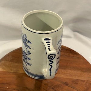 May include: Cream ceramic mug with blue floral patterns and a blue handle. The mug is cylindrical with a wide opening. The handle is decorated with blue lines and swirls. The mug has the text "Yacht Club" in blue.