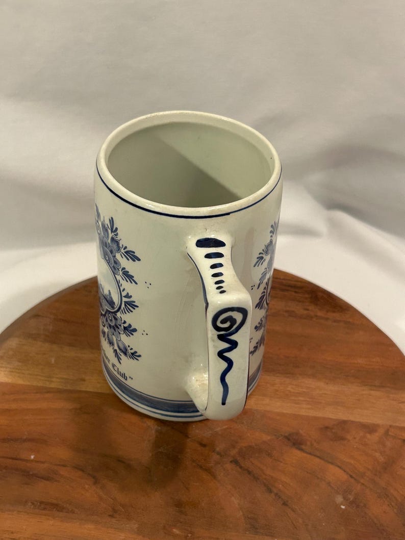 May include: Cream ceramic mug with blue floral patterns and a blue handle. The mug is cylindrical with a wide opening. The handle is decorated with blue lines and swirls. The mug has the text "Yacht Club" in blue.