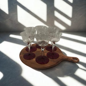 May include: A set of six clear crystal glasses with red bases, arranged on a wooden serving board. The glasses have etched floral designs. The board has a handle. The scene is lit by sunlight, casting shadows on the surface.