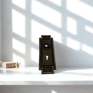 以下が含まれることがあります： キーホールとネジ穴が付いた、ヴィンテージのブロンズトーンのドアプレート。長方形のプレートには装飾的な上部と下部があります。小さな金色の物体がプレートの左側にあります。アイテムは、窓の光の影がある白い棚にあります。
