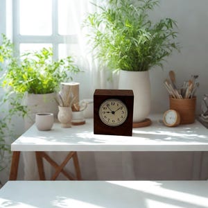May include: A dark brown, square-shaped clock with a cream-colored face and black numerals sits on a white table. The clock has a classic design, with a round clock face. The background includes green plants in white pots and a window.