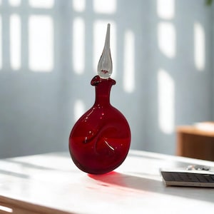 Puede incluir: Una botella de vidrio rojo con un tapón de vidrio transparente. La botella tiene una forma redondeada con un tapón largo y puntiagudo. La botella está sobre una superficie blanca.