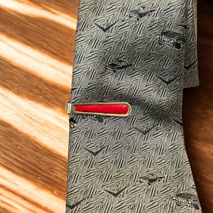 May include: A silver-toned tie bar with a bright red rectangular inlay attached to a grey necktie. The tie features a repeating pattern of small black vintage cars and chevron shapes woven into a textured, light grey fabric background.