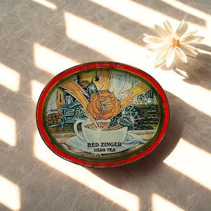 Puede incluir: Lata ovalada con borde rojo, diseño floral y el texto "Red Zinger Herb Tea". También se ve una margarita blanca y sombras de la luz solar.