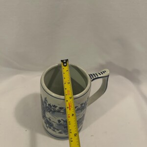 May include: A blue and white ceramic mug with a handle. The mug has a decorative landscape design and is approximately 5 inches tall. The mug is being measured with a yellow tape measure.