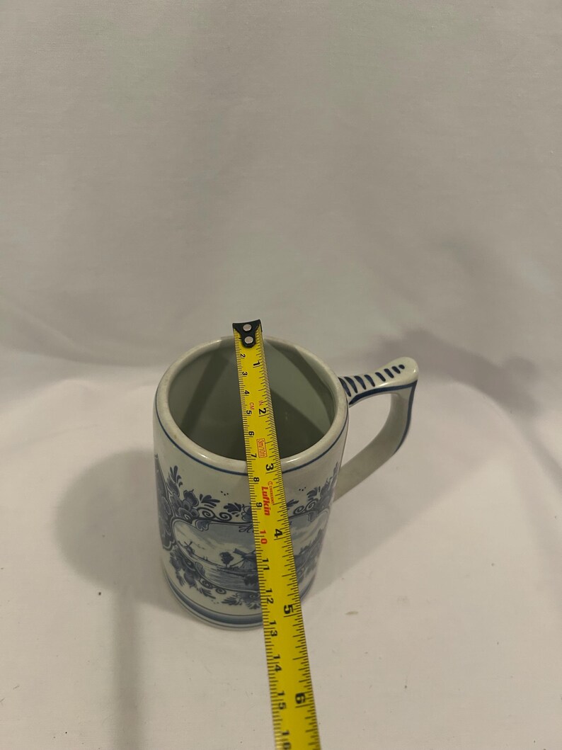 May include: A blue and white ceramic mug with a handle. The mug has a decorative landscape design and is approximately 5 inches tall. The mug is being measured with a yellow tape measure.