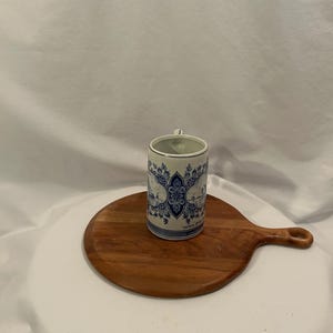 May include: A cylindrical ceramic mug with a blue and white floral pattern, complete with a small handle. It rests on a round wooden chopping board with a handle. The backdrop is a white cloth.