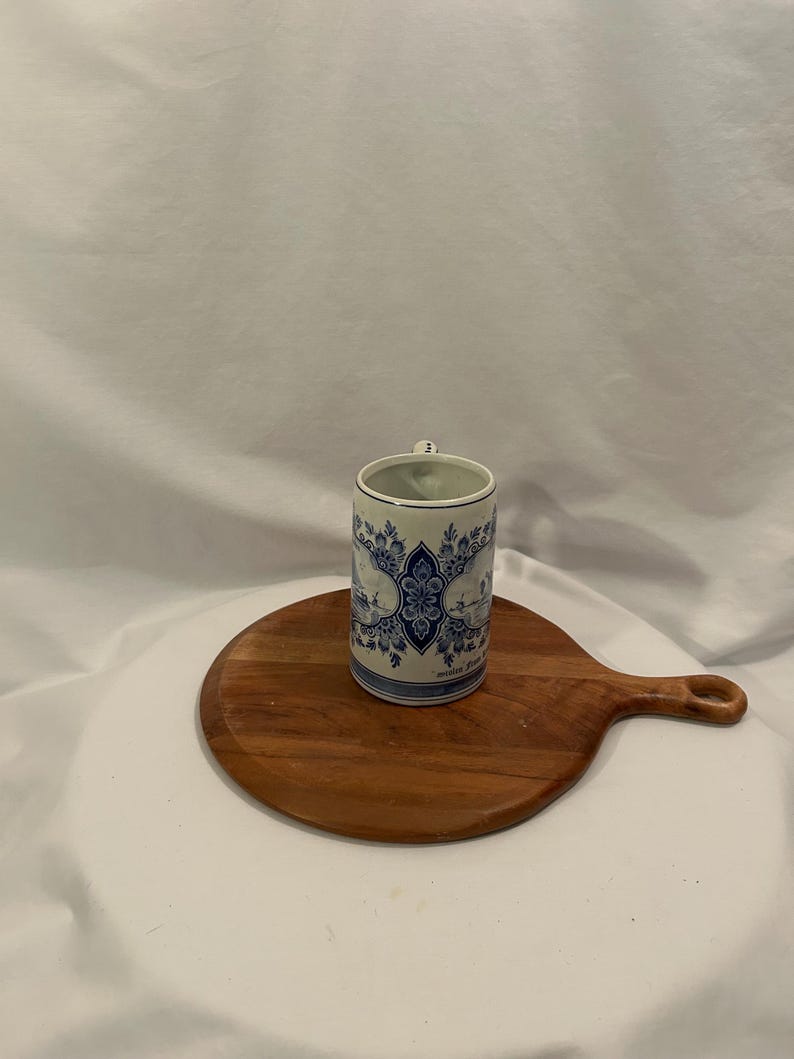 May include: A cylindrical ceramic mug with a blue and white floral pattern, complete with a small handle. It rests on a round wooden chopping board with a handle. The backdrop is a white cloth.