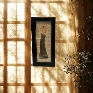 May include: A framed art print featuring a vintage illustration of a woman in a long dress with a low back. The artwork is set against a backdrop of aged book pages, framed in a black rectangular frame. The artwork is displayed near a window.