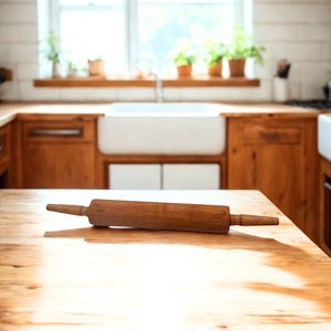 May include: A wooden rolling pin with dark brown handles rests on a light brown wooden countertop. The kitchen setting includes a white farmhouse sink, wooden cabinets, and a window with plants. The rolling pin is a classic kitchen tool.