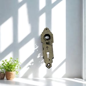 May include: An antique, ornate door plate with a keyhole and a circular opening. The metal plate has a distressed, aged appearance with a faded, off-white finish. The door plate is mounted on a white wall, with shadows from a window.