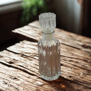 May include: A clear glass decanter featuring a vertical ribbed texture on the body and a matching faceted glass stopper. The vessel has a cylindrical shape with a slightly rounded base and a narrow neck designed to hold the removable glass lid securely.