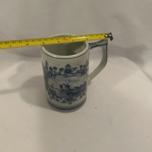 May include: A white and blue ceramic mug with a handle. The mug features a Dutch windmill scene and the word "Heineken". The mug is approximately 4 inches tall.