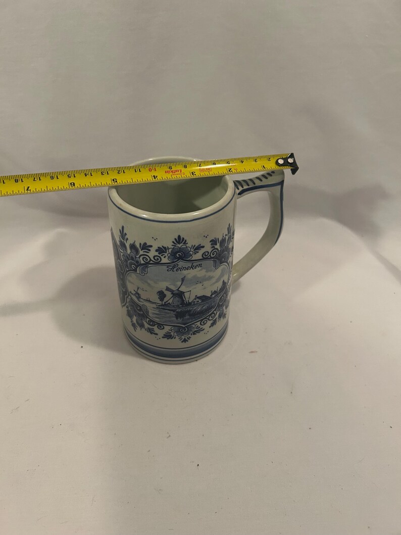 May include: A white and blue ceramic mug with a handle. The mug features a Dutch windmill scene and the word "Heineken". The mug is approximately 4 inches tall.
