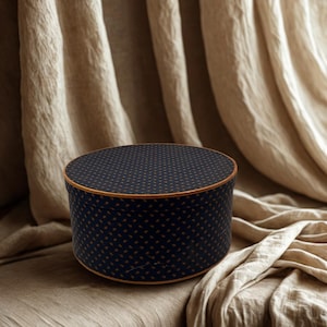 May include: A round, navy blue box with a repeating gold pattern and a gold trim. The box is sitting on a beige surface with a beige draped background.