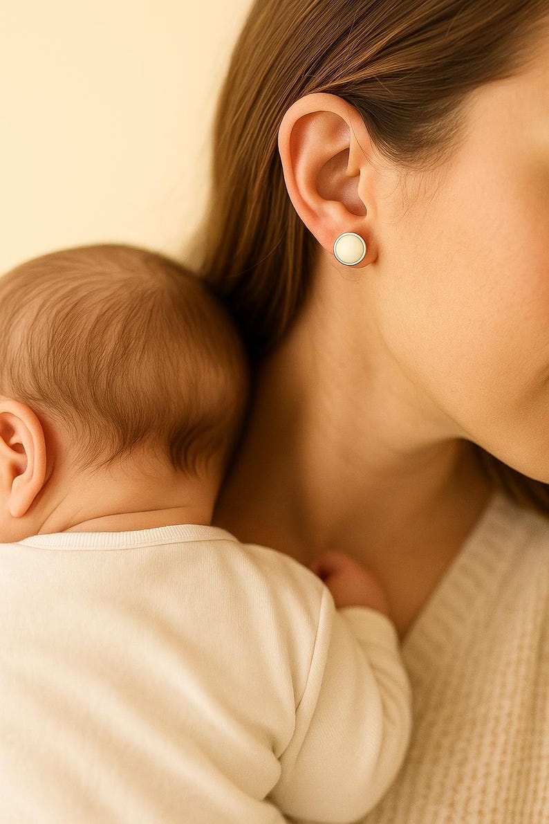 May include: A close-up of a woman's ear with a white, round stud earring. A baby is resting on the woman's shoulder. The baby is wearing a white onesie. The background is a soft, warm color.