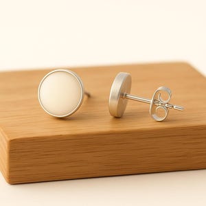 May include: A pair of silver stud earrings with a circular, off-white center. The earrings are displayed on a wooden surface. The earrings have a simple, minimalist design.