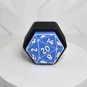May include: A blue and white twenty-sided die, with white numbers, sits inside a black hexagonal frame. The die is on a white cylindrical platform against a white geometric background. This item is likely for tabletop gaming.