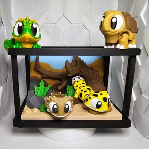 May include: A black framed terrarium with cartoon reptile figurines. The figurines include a green and yellow gecko, a brown turtle, and two yellow and black spotted geckos. The terrarium has a desert scene backdrop.