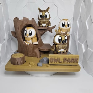May include: A 3D-printed diorama featuring four owl figurines in a woodland setting. The scene includes a tree, a stump, rocks, and a sign that reads "OWL PARK". The owls are in shades of brown and tan.