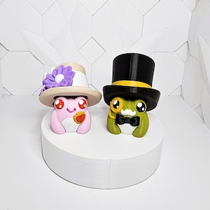 May include: Two whimsical frog figurines. One is pink with a hat and a flower, the other is green with a top hat and bow tie. Both are on a white circular platform, set against a white geometric background.