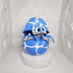 May include: A blue and white dragon figurine emerging from a matching egg. The egg is patterned with white spots. The dragon has tan wings and a friendly expression. The figurine is displayed on a white pedestal.