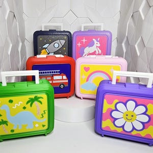 May include: A collection of six small rectangular plastic cases with white handles. The cases feature colorful designs including a blue dinosaur, a red fire truck, a white rocket, a purple unicorn, a rainbow, and a yellow daisy with a smiley face.