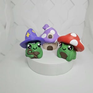 May include: Three whimsical frog figurines. One frog wears a purple wizard hat with a yellow star, another has a red mushroom cap with white spots, and the third has a purple mushroom house. All are green with brown bellies.