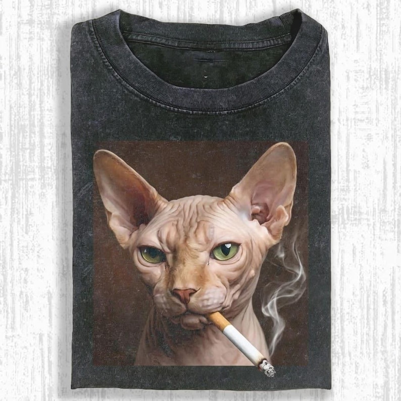 May include: Dark gray t-shirt featuring a portrait of a Sphynx cat with green eyes smoking a cigarette. The cat is centered on a brown background, with smoke rising from the cigarette. The shirt has a crew neck.