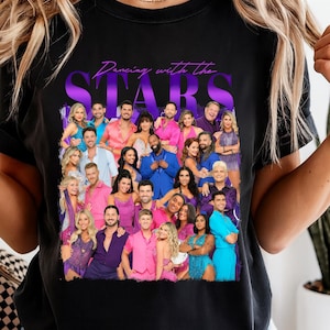 Può includere: T-shirt nero con una foto di gruppo di persone in abiti colorati. Il testo "Dancing with the Stars" è stampato in viola e bianco. La maglietta è realizzata in un materiale morbido.