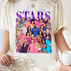 May include: Ivory-colored t-shirt featuring a group photo of people in colorful outfits. The words "Dancing with the STARS" are printed in purple above the photo. The word "IVORY" is printed in red at the bottom left.