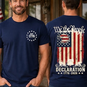 May include: Navy blue t-shirts with patriotic designs. One shirt features "EST 1776" and stars. The other displays an American flag with an eagle, the words "We the People" and "Declaration 1776-2026".