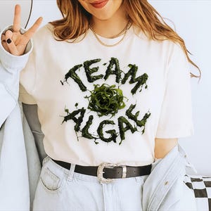 May include: A cream-colored short-sleeve t-shirt featuring the text "TEAM ALGAE" in a dark green, textured, slime-like font. A central circular graphic of dark green, swirling algae sits between the two words. The shirt is worn with light blue jeans.