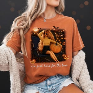 Puede incluir: Una camiseta naranja de manga corta con el gráfico de una muñeca de pelo rubio largo, con un suéter amarillo y falda negra, sentada en una silla con una calabaza. El texto "i'm just here for the boos" está impreso debajo del gráfico.
