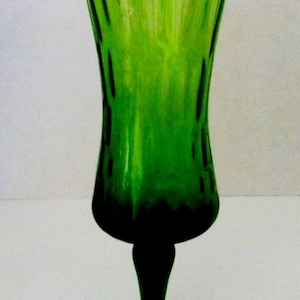 May include: A tall, green glass vase with a flared, scalloped rim. The vase has a dark green base and stem, and a round base. The body of the vase has vertical lines and a textured appearance. The vase is approximately 10 inches tall.
