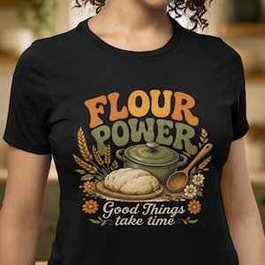 Op de afbeelding: Een zwart t-shirt met een grafische print met de tekst "FLOUR POWER" en "Good Things take time". Het ontwerp bevat een groene braadpan, een brood op een houten plank, een houten lepel, korenaren en oranje en witte bloemen.