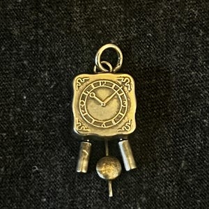 May include: A small, antique-style clock pendant with a square face and decorative scrollwork. The clock face shows the time as approximately 2:10. The pendant has a loop at the top for hanging and a pendulum design at the bottom.