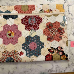 May include: A patchwork pouch with a cream zip. The pouch is made from hexagonal fabric pieces in cream, pink, red, and blue. The design features floral and patterned motifs. The label reads "Dragonfly Studio".