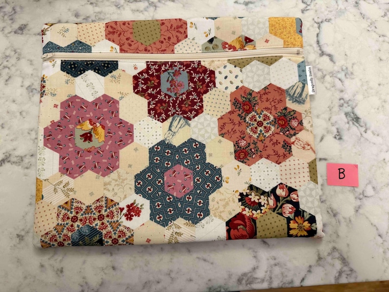 May include: A patchwork pouch with a cream zip. The pouch is made from hexagonal fabric pieces in cream, pink, red, and blue. The design features floral and patterned motifs. The label reads "Dragonfly Studio".