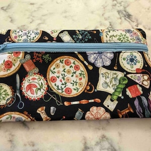Op de afbeelding: Een rechthoekig stoffen etui met rits op een zwarte achtergrond, bedrukt met kleurrijke borduurringen, garenklosjes, scharen en speldenkussens. Een lichtblauwe rits loopt over de bovenkant en een wit label bevat de tekst Dragonfly Realm.