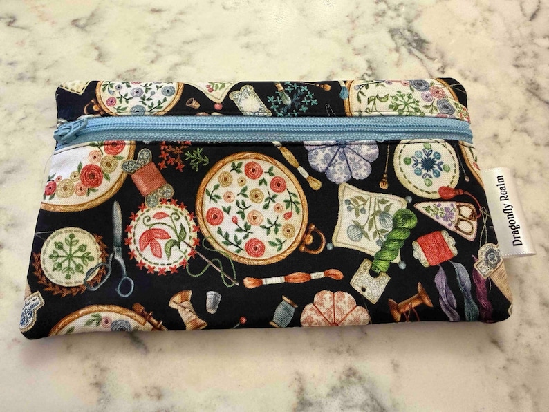 Op de afbeelding: Een rechthoekig stoffen etui met rits op een zwarte achtergrond, bedrukt met kleurrijke borduurringen, garenklosjes, scharen en speldenkussens. Een lichtblauwe rits loopt over de bovenkant en een wit label bevat de tekst Dragonfly Realm.