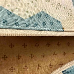 May include: Close-up of a fabric item with a cream-coloured zip. The fabric features a patchwork design with blue, cream, and floral patterns. The interior lining is a light tan colour with a repeating cross pattern.