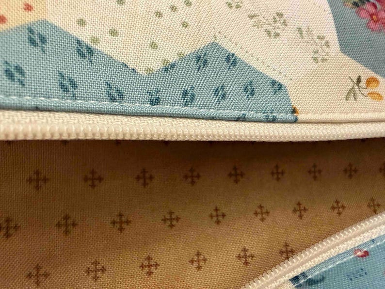 May include: Close-up of a fabric item with a cream-coloured zip. The fabric features a patchwork design with blue, cream, and floral patterns. The interior lining is a light tan colour with a repeating cross pattern.